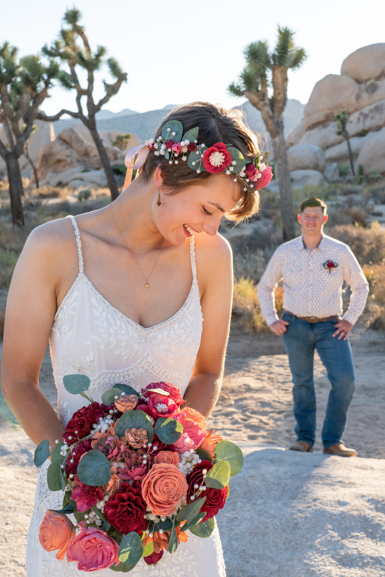Boho bride poses with headband and flowers smiling and laughing while groom stands in background with hands on hips Hidden Valley Joshua Tree California