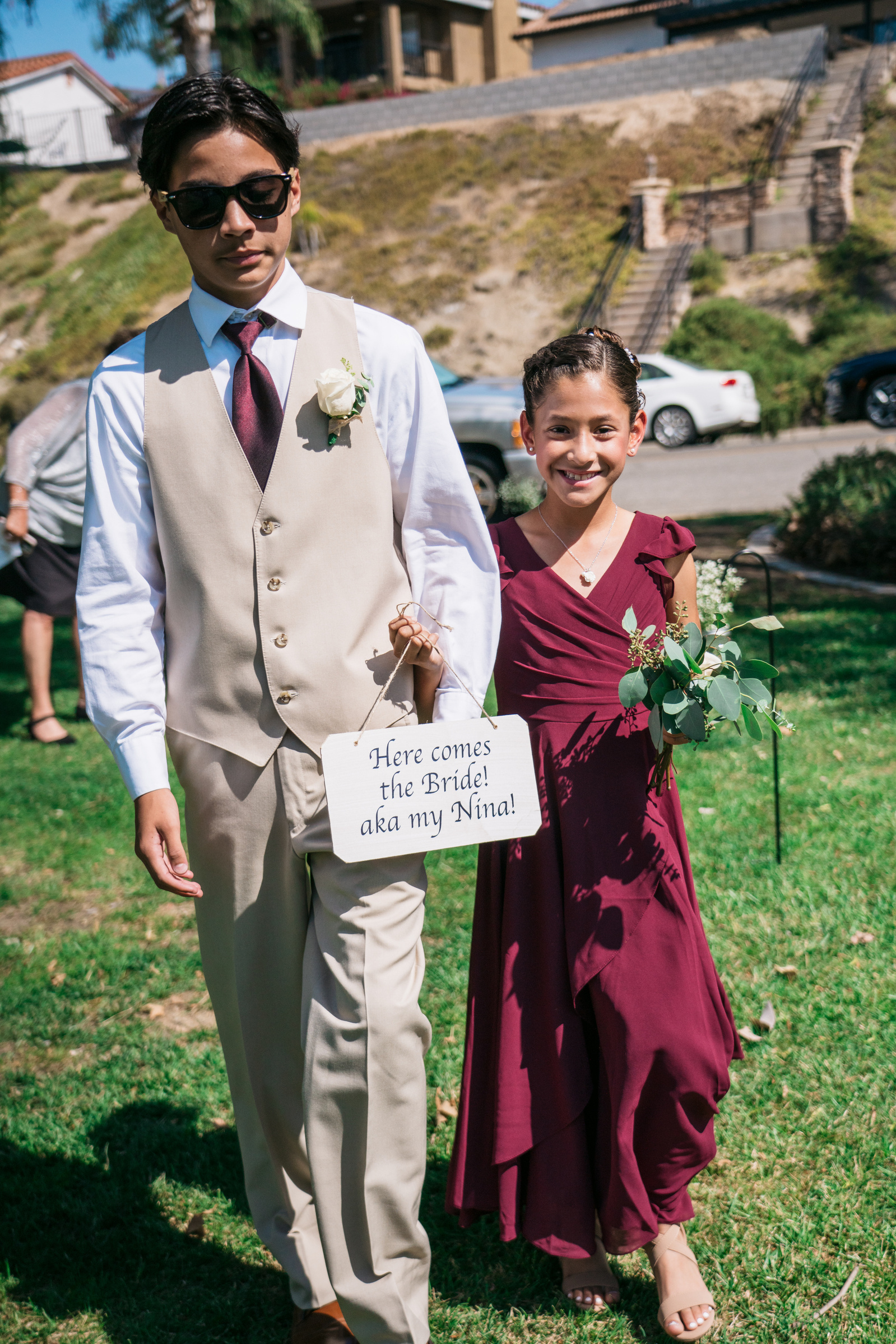 junior groomsman and bridesmaid arm in arm holding sign that reads "here comes the bride aka my nina"