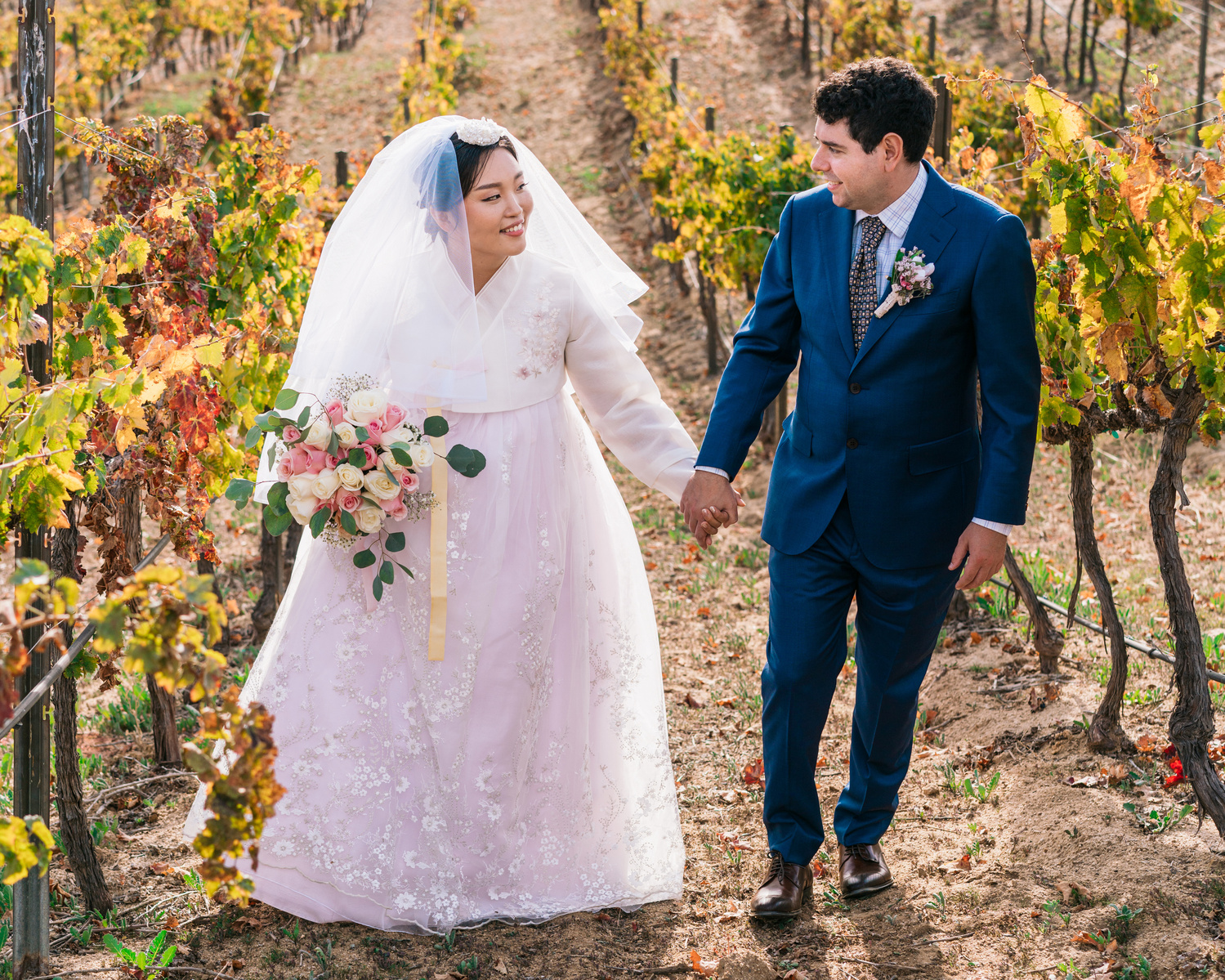 korean bride in her hanbok and groom walk among the vines in the vineyard hand in hand staring into each others eyes