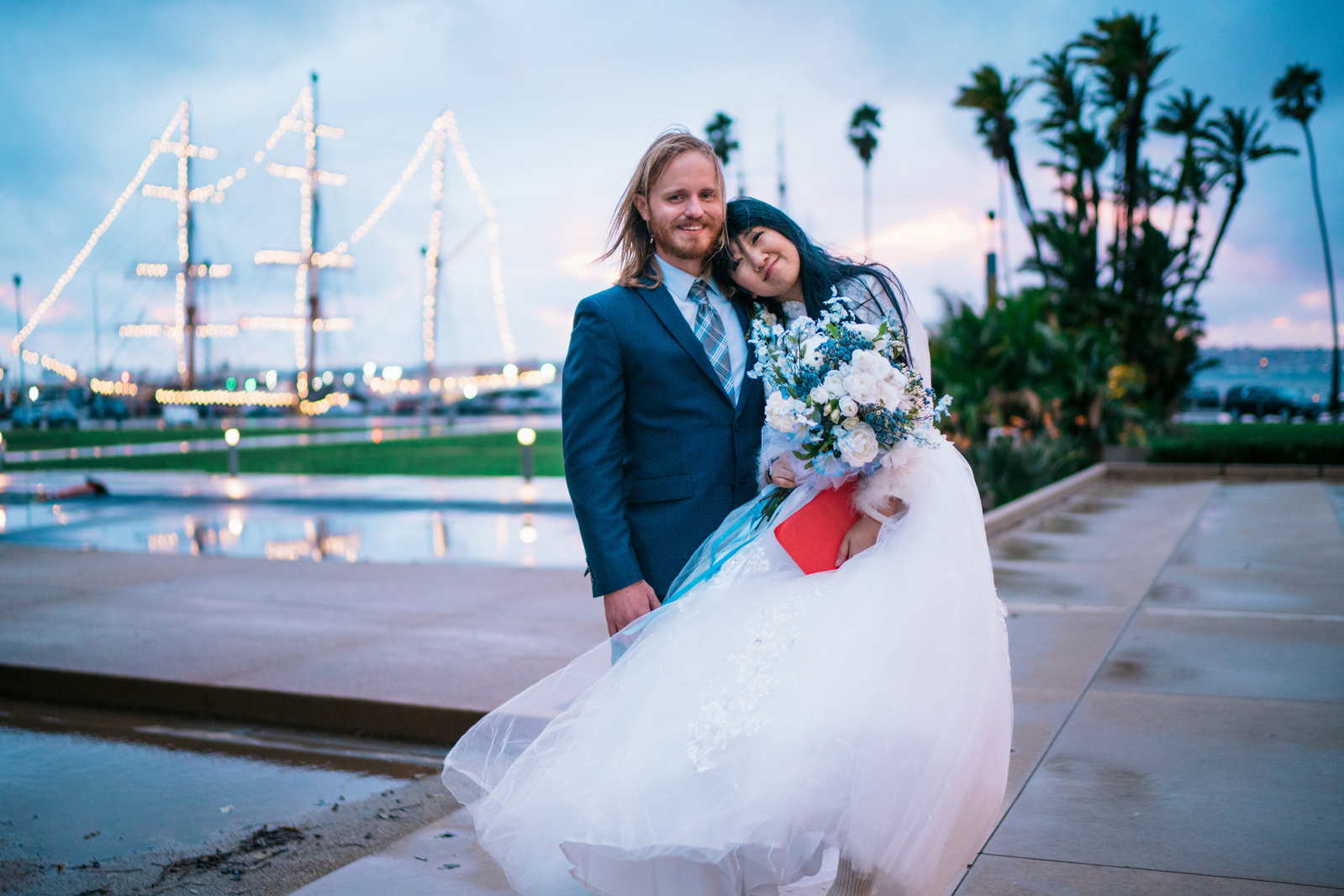 On a rainy day husband and wife pose for a portrait in front a ship and palm trees after their wedding at San Diego City Hall
