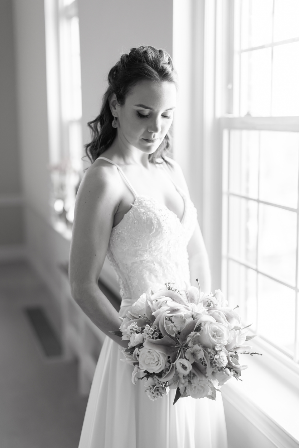 A bride stood next to a window looking down at her wedding bouquet, with sunlight illuminating.  