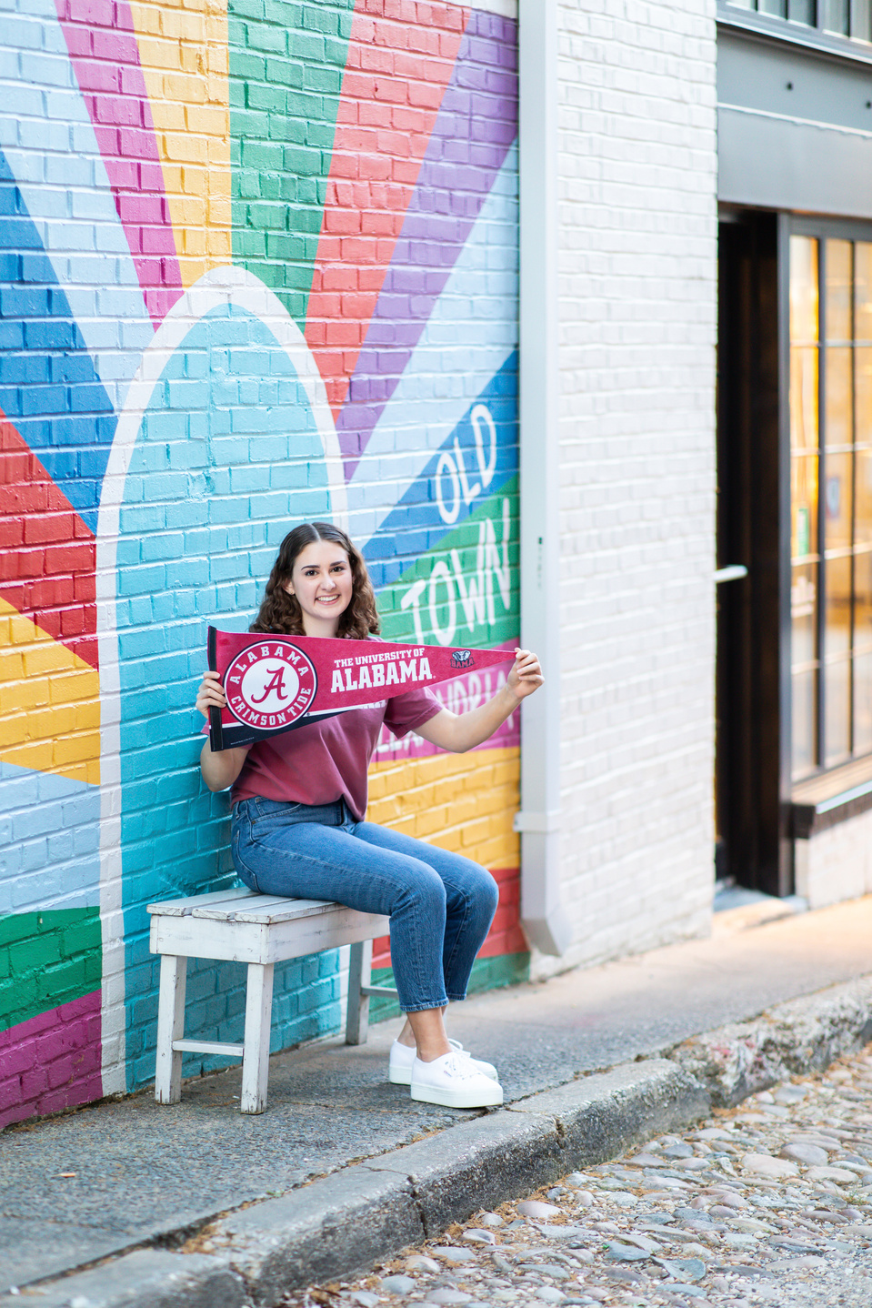 Girl sat on a bench holding a sign reading "University of Alabama" with a rainbow background and the words "Old Town Alexandria" .