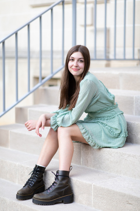 A natural light photograph of a teenage girl sat on some steps, wearing a green dress and Dr Martin black lace up boots.
