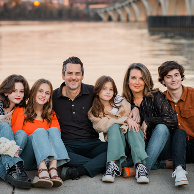 A family sat next to the Potomac river with a bridge in the background at sunset.