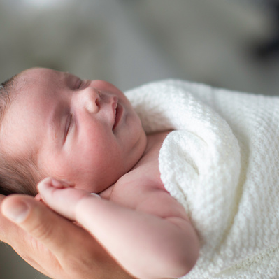 A newborn baby wrapped in a white blanket, held in the hand of her father.