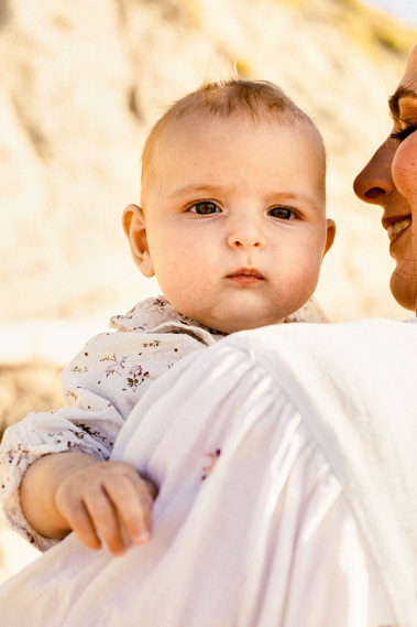 Un bébé qui regarde la caméra et qui est dans les bras de sa mère sur la plage.