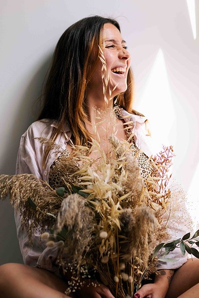 Une femme rit aux éclats en tenant un bouquet de fleurs séchées lors d'une séance photo grossesse à domicile à Toulouse.