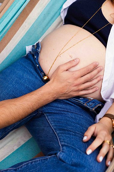 Le ventre d'une femme enceinte sur un hamac coloré et avec la main du futur papa lors d'une séance photo maternité à Toulouse