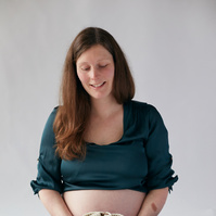 Maternity photograph showing mother-to-be holding baby booties taken in a photography studio against plain white background.