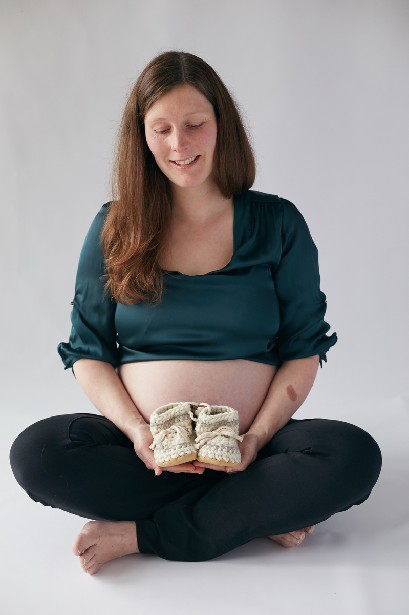 Maternity photograph showing mother-to-be holding baby booties taken in a photography studio against plain white background.