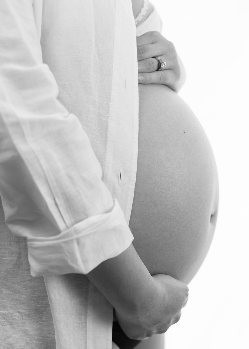 Professional Maternity photograph of a womans pregnant belly, wearing white shirt against a white backdrop.
