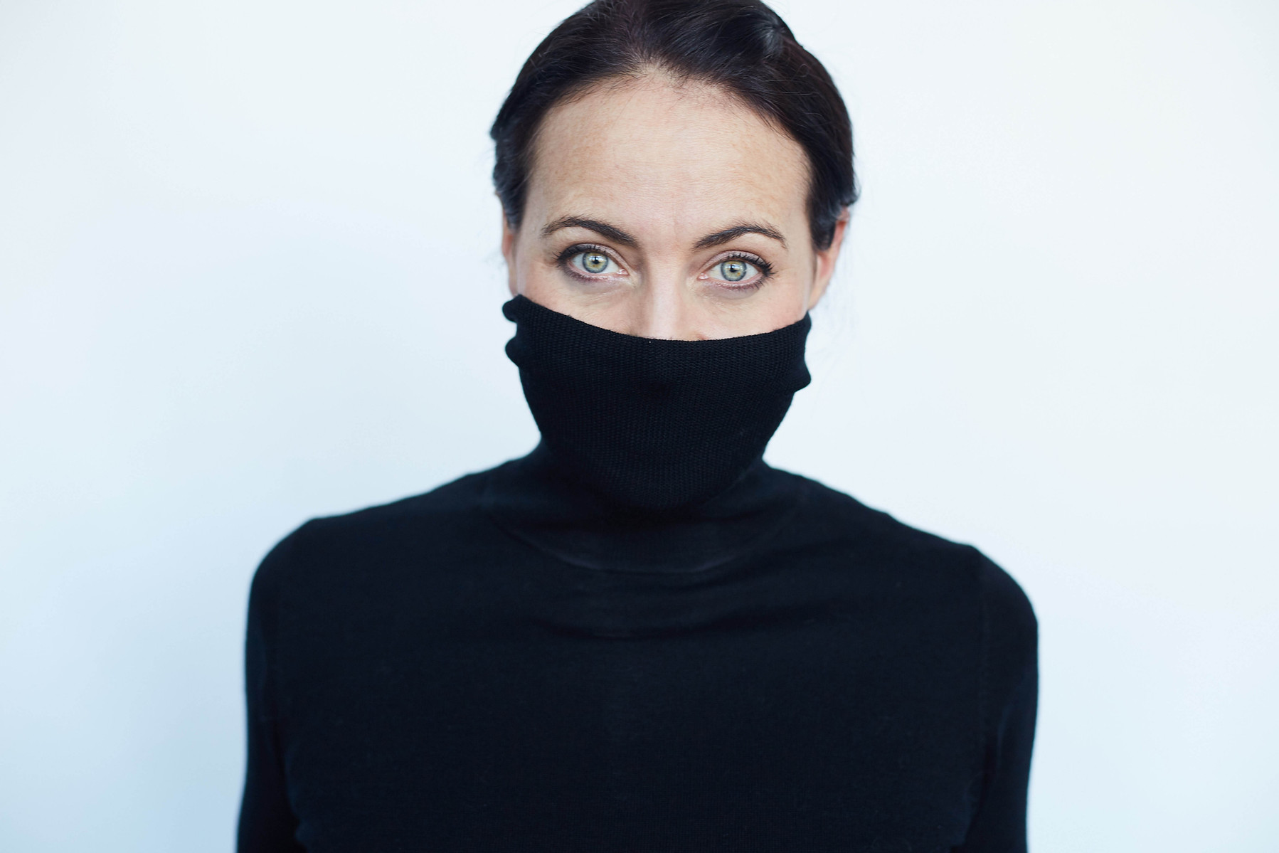 Contemporary portrait of a woman wearing black polo neck turtle neck jumper pulled over her mouth, with striking blue eyes. 