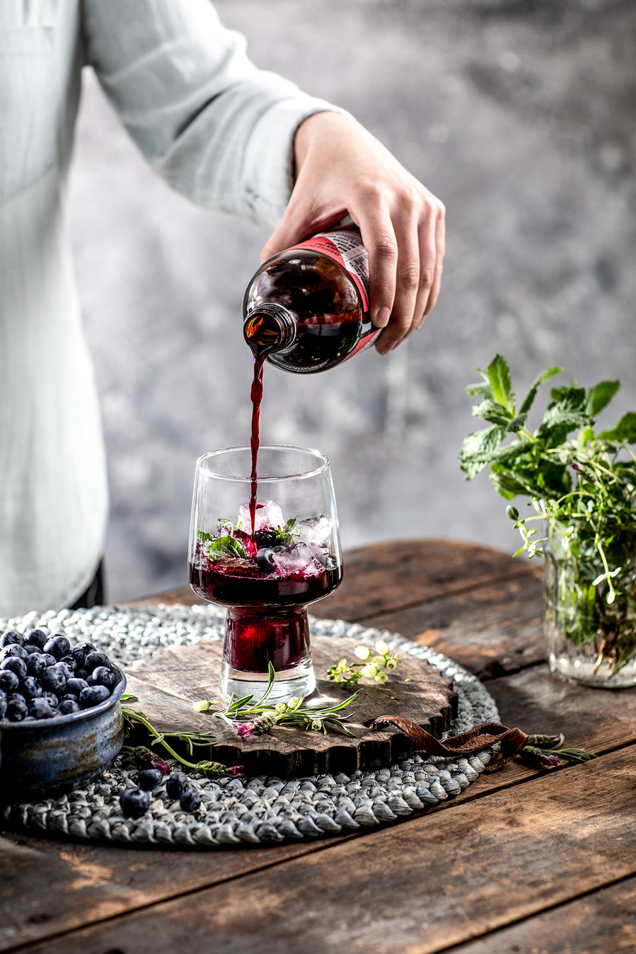 A person pouring wine into a blueberry sangria thick-stemmed glass on a rustic table with a mint plant a a dish of fresh blueberries. 