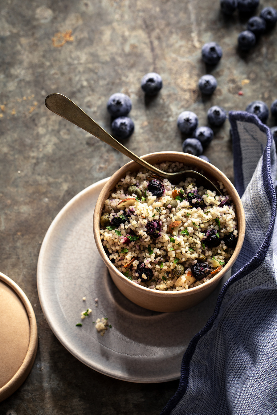 Blueberry quinoa salad in a small croc with a spoon. Sun with moody shadows with stoneware props and more blueberries on surface. 
