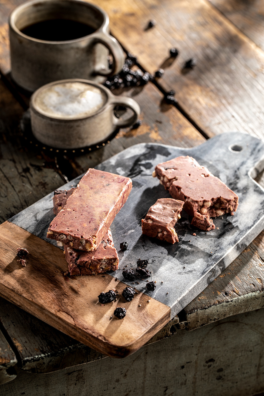 Blueberry and chocolate granola bars on a marble and wood cutting board with dried blueberries. All on a wooden table with stoneware mugs with foamy espresso. Moody branding photography for the blueberry council. 