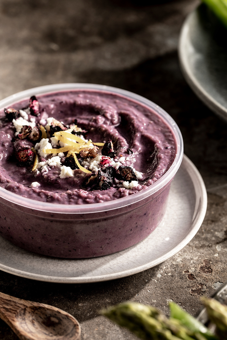 A plastic container of blueberry hummus with toppings on a concrete surface with other plates and a wooden spoon. 