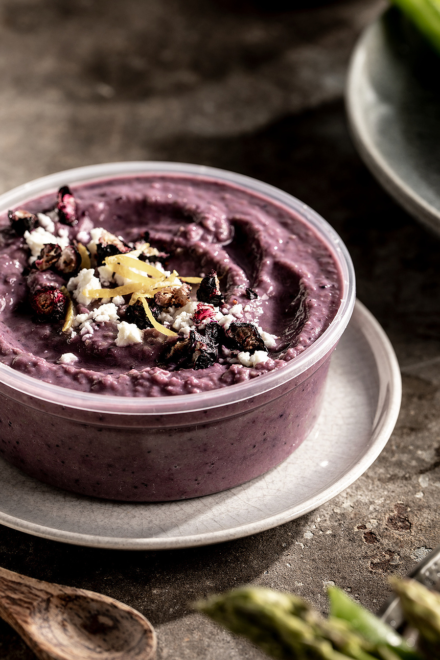 A plastic container of blueberry hummus with toppings on a concrete surface with other plates and a wooden spoon. 