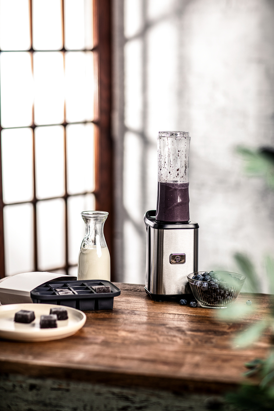 A small blender on a wooden counter in front of a patio door with blueberry smoothie inside. Milk and tray of frozen portioned blueberries sits off to the side. Branding photography for the blueberry council. 