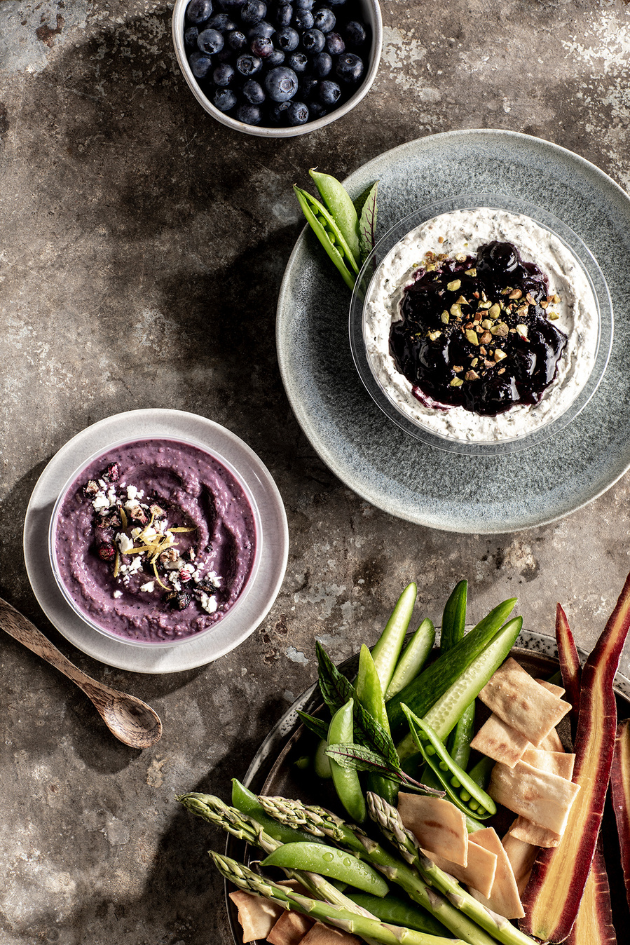 Overhead shot of two types of blueberry hummus in separate bowls, with a tray of veggies and cheese for dipping and a bowl of fresh blueberries. Images of blueberries for the blueberry council. 