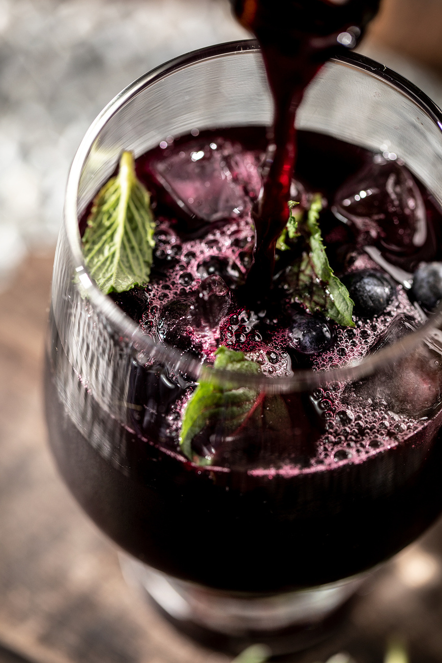 Looking into a glass of blueberry sangria with fresh mint leaves. A bottle of wine out of frame is pouring into the glass. branding photography for the blueberry council.  