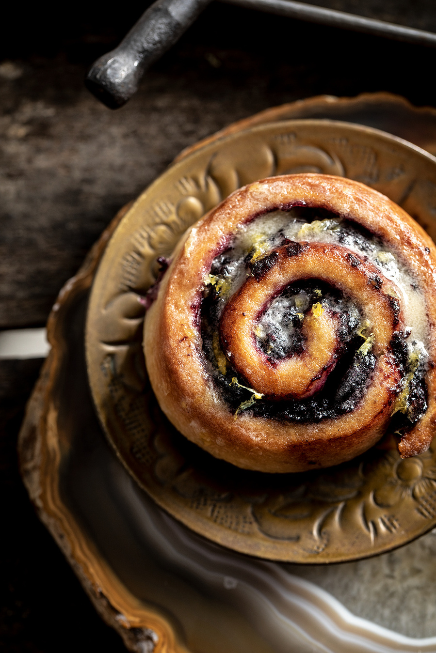 Overhead shot of jumbo blueberry cinnamon roll on a bronze plate with moody backdrop. Branding photography for the blueberry council. 
