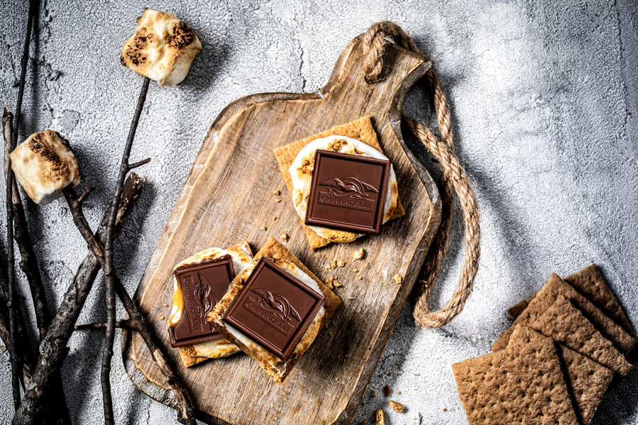 Overhead Ghirardelli chocolate photography with chocolate squares on open faced s'mores, more graham crackers and toasted marshmallows on sticks.