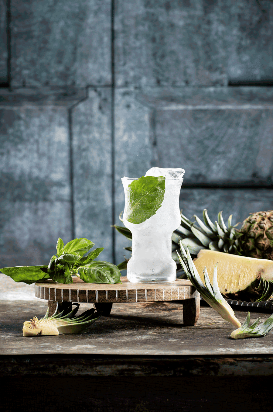 Drink photographer gif of a hand pouring a can of Press Seltzer into a wide stem glass with ice and a basil leaf. Sliced pineapple in the background with dark blue backdrop.  