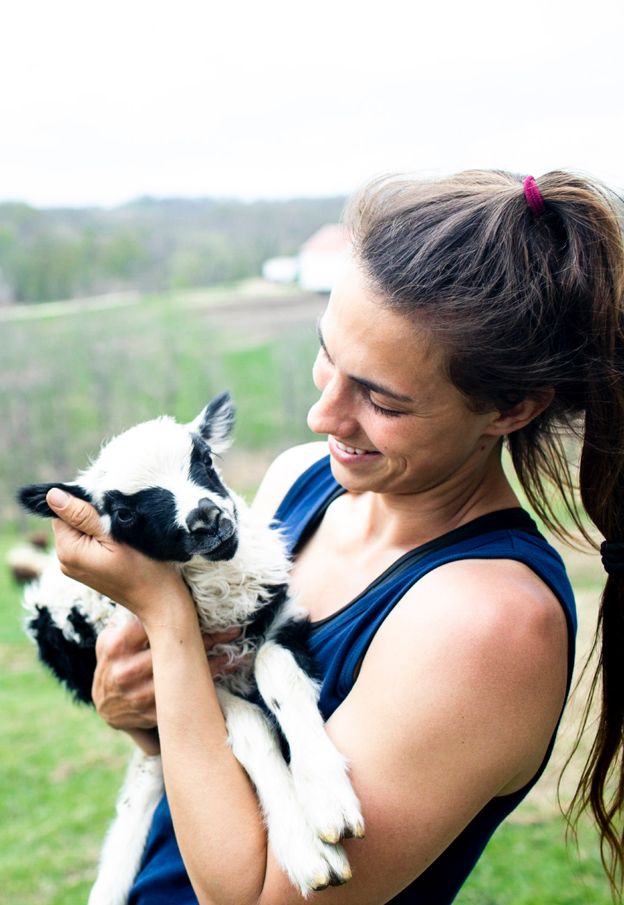 Portrait of a female farmer holds a baby lamb in her arms and is smiling at it. Branding photography for agriculture pork and beef farm. 