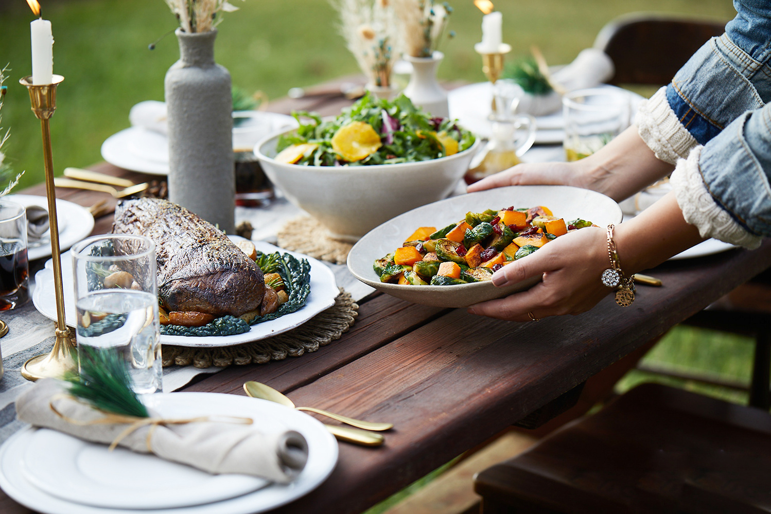 Beautiful rustic glam table setting on outdoor picnic table. Arms of woman in jean jacket sets a dish on the table. 