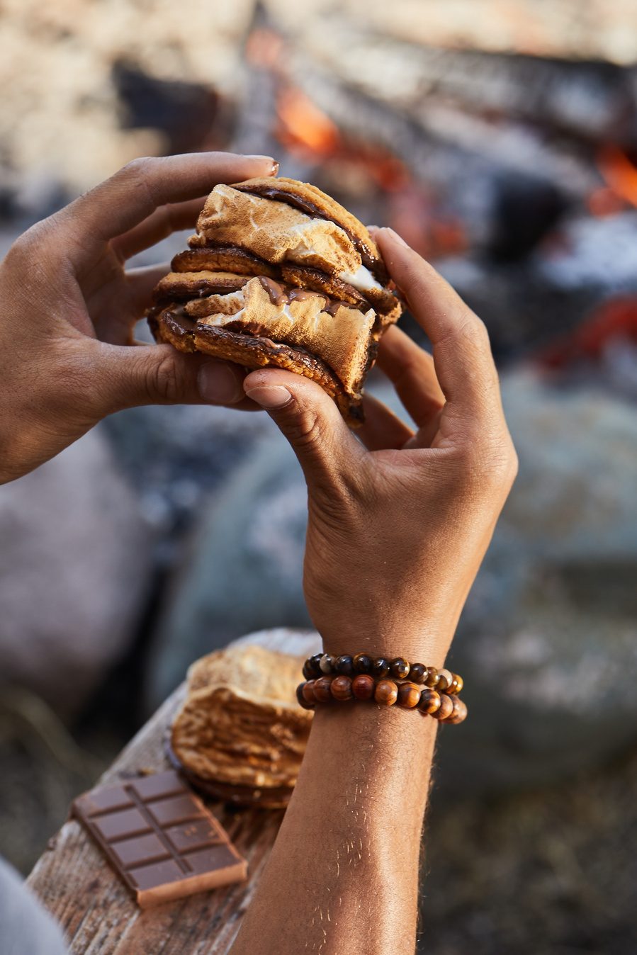 A man's hands hold a large smore in front of an outdoor fire pit. Lifestyle food photography for The Buckle