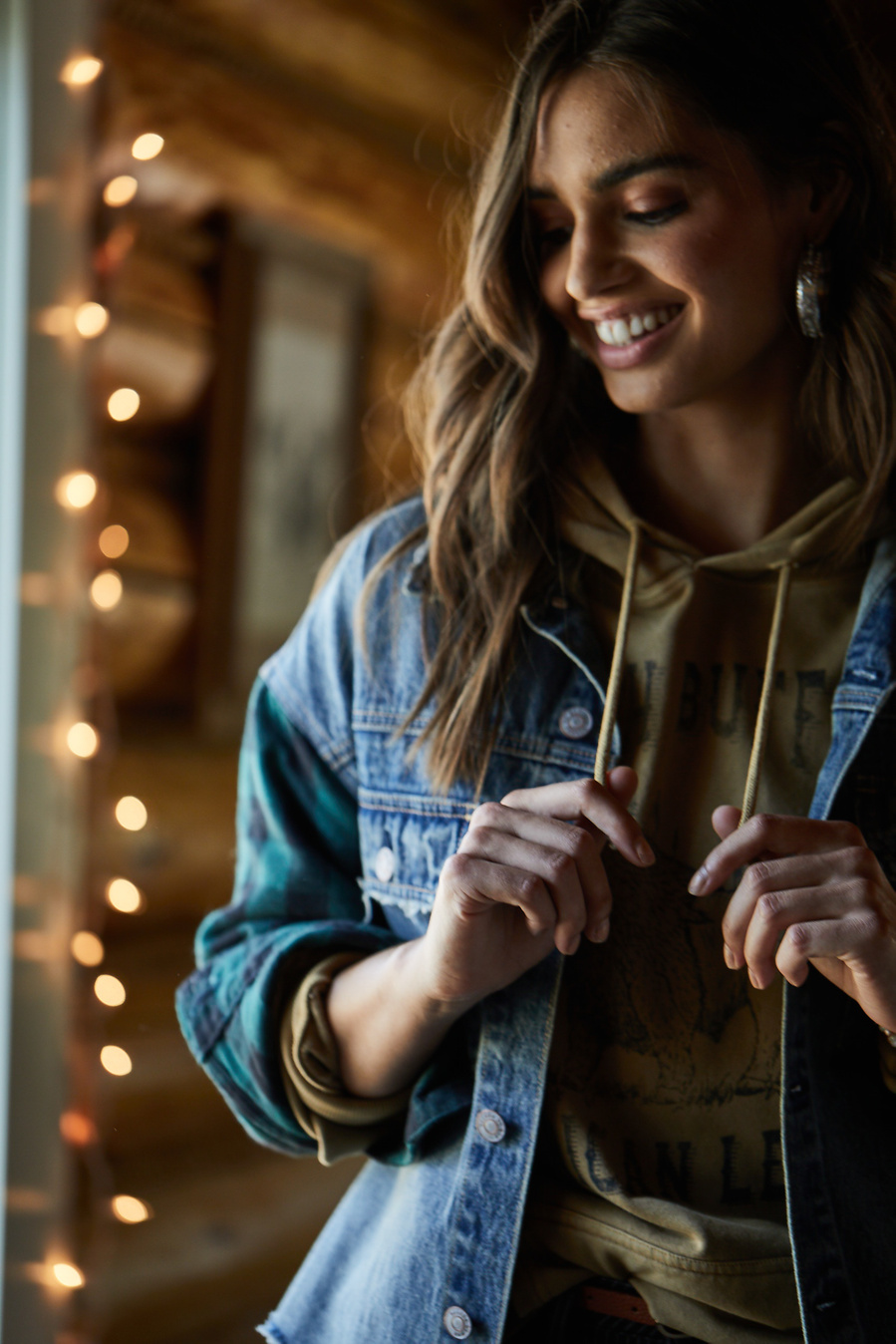 Female fashion model poses in sweatshirt and jean jacket with sleeves rolled up and hands holding the sweatshirt ties in a winter cabin. Fashion lifestyle photography for The Buckle