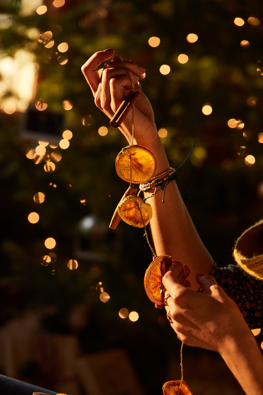 Hands holding up a string of orange slices and cinnamon in front of a lit Christmas tree glowing in the light of the sunset. Lifestyle food photography for The Buckle. 