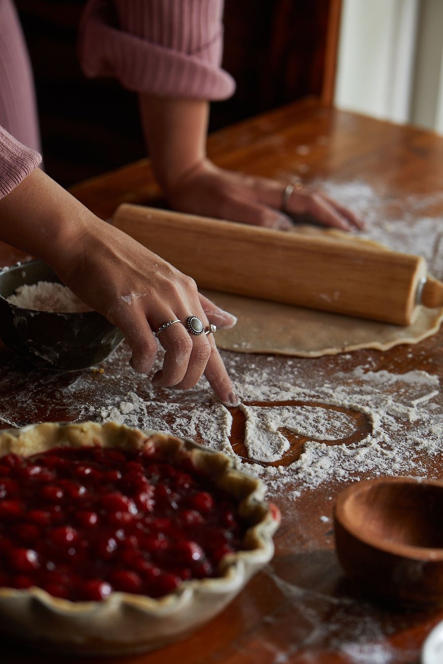 Woman in pink sweatsuit draws a heart in the flour spread on the table with a rolling pin and pie crust in the background and cherry pie in the foreground. Fashion and Lifestyle food photography for The Buckle
