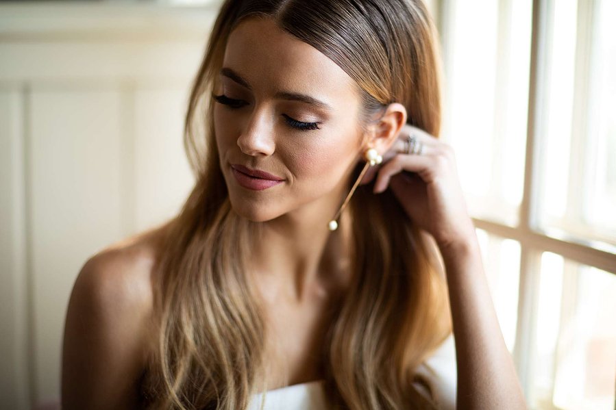 bride near a window looking away from the light with hand in hair showing gold earrings