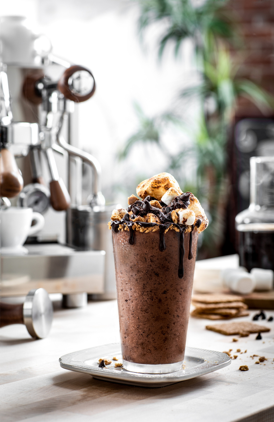 Frozen chocolate s'more coffee drink in tall glass on a saucer with a graham cracker rim, toasted marshmallows and dripping chocolate sauce. Bright and moody chocolate photographer. 