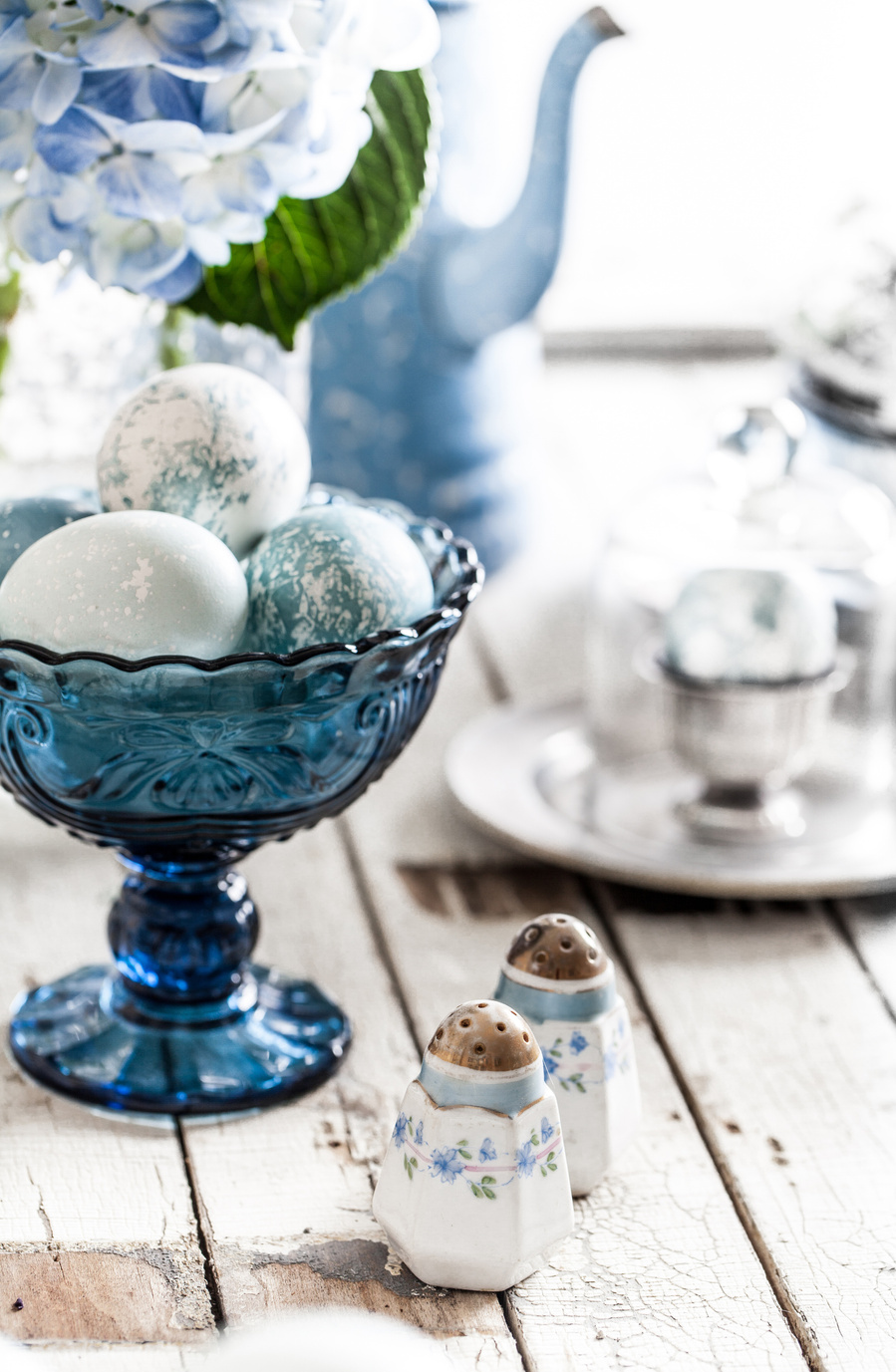 Styled blue glass dish with blue and white dyed eggs on a white wood surface with blue hydrangeas, a tea set, and salt and pepper on the table. Final result of diy dyed eggs photography 