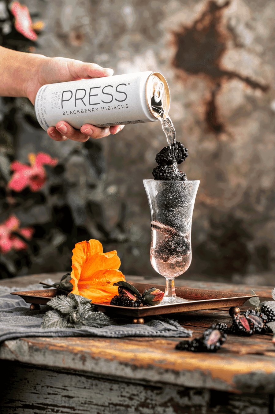 drink photography gif of can of press seltzer pouring into a stemmed glass of ice and blackberries with an orange hibiscus flower on the surface. 