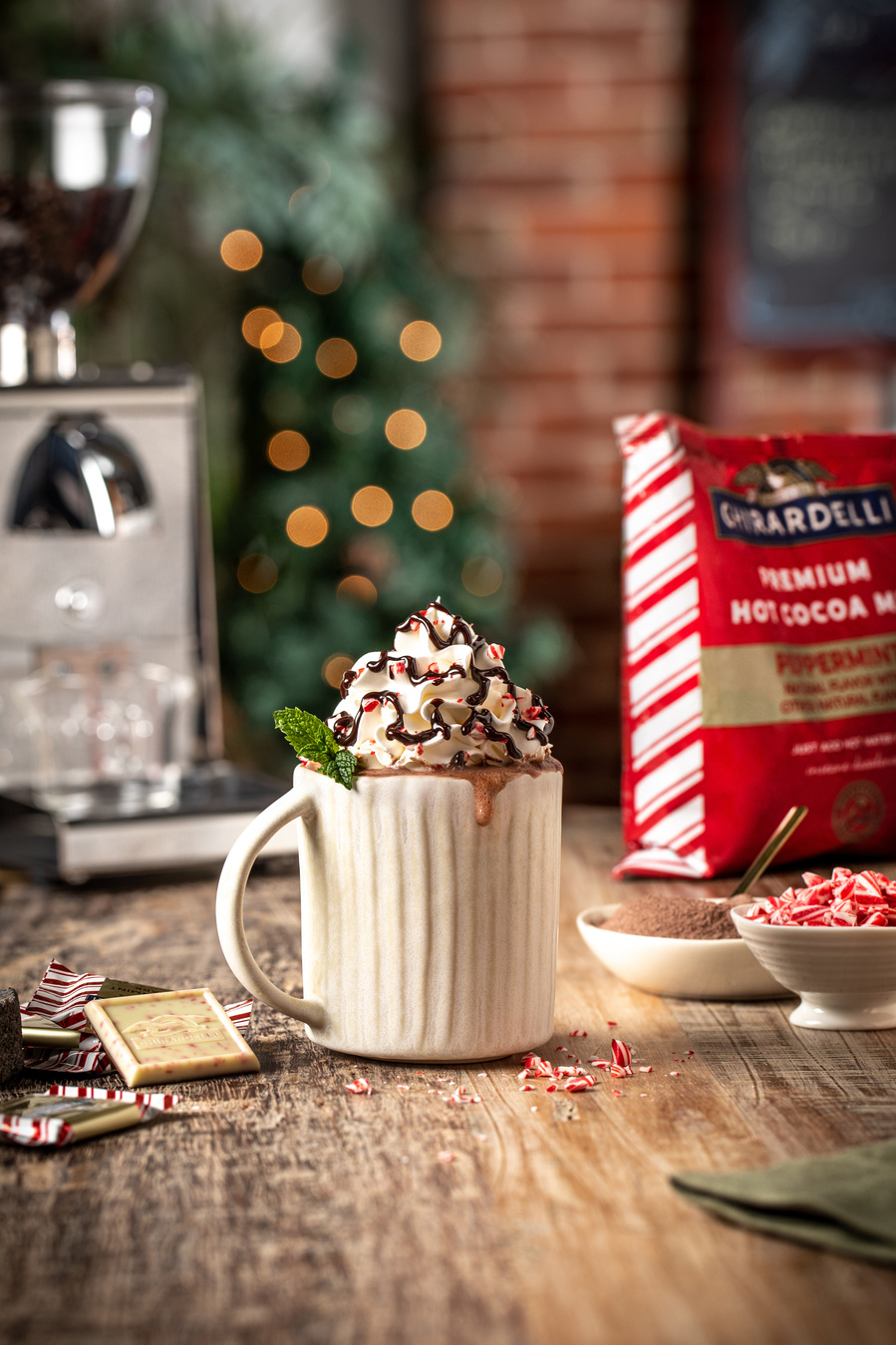 A white mug with a drip of hot chocolate down the side. Topped with whipped cream, chocolate sauce, and crushed peppermint. Espresso machine a Ghirardelli packaging in the back and unwrapped chocolate in front. 