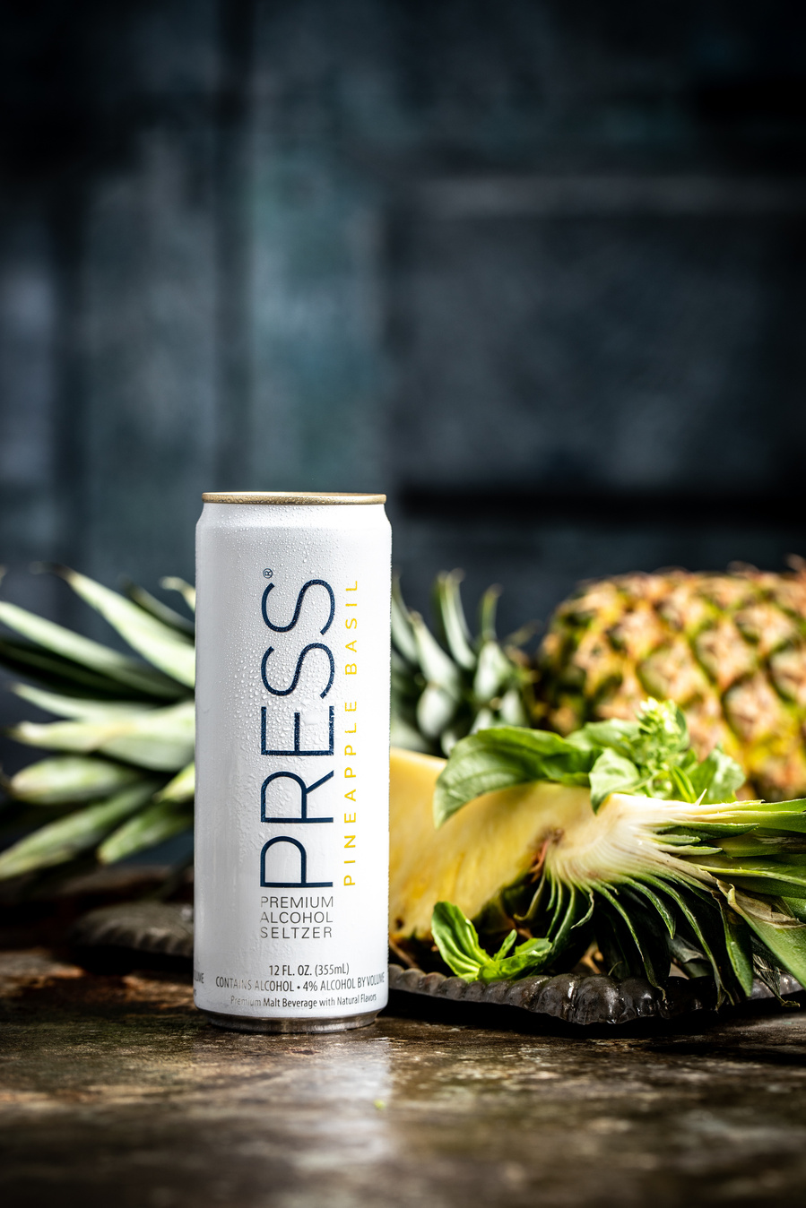 Pineapple basil can of Press Seltzer with sliced and whole pineapples behind. Drink photography with dappled light and dark backdrop. 