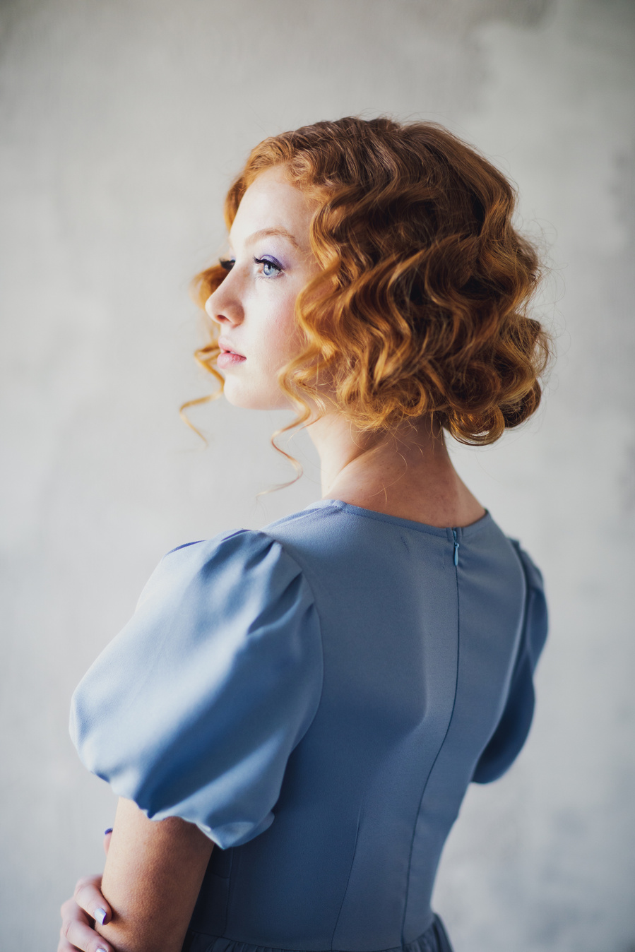Model with red cropped curls in powder blue dress looking into the light with her back to the camera. Lifestyle editorial photography of feminine tea party food photo shoot. 