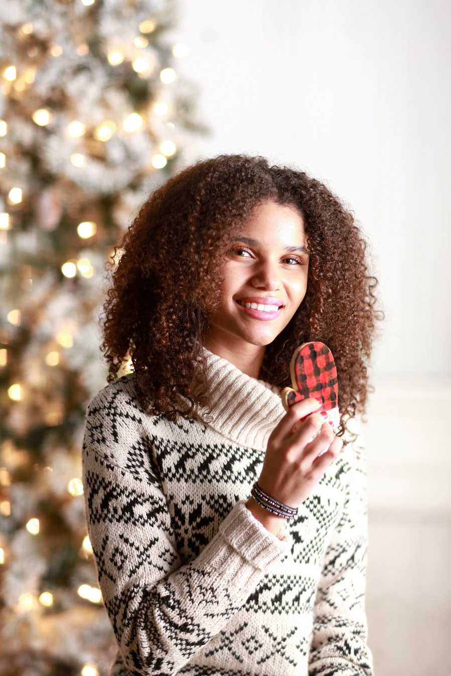 Model with curly brown hair in a striped white and black faire isle sweater with a cowl neck sitting in front of a Christmas tree and holding a buffalo plaid mitten cookie as if to eat it. Part of a brand photography campaign for Maurices
