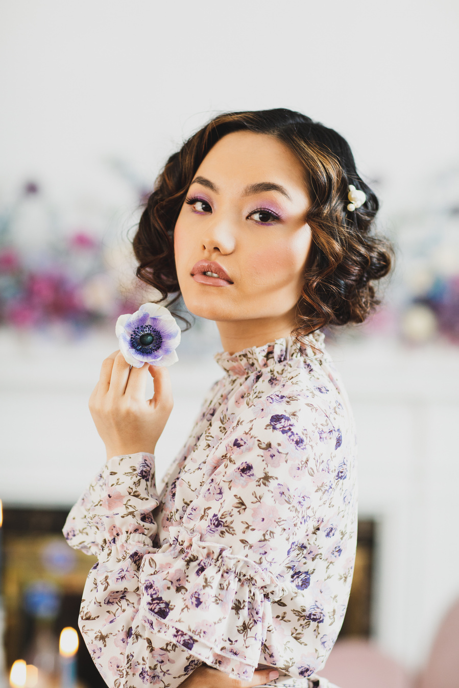 Editorial photography of model with cropped brown curls and a floral blouse looking at the camera, holding a purple flower near her face at a Bridgeton style dessert photography tea party. 
