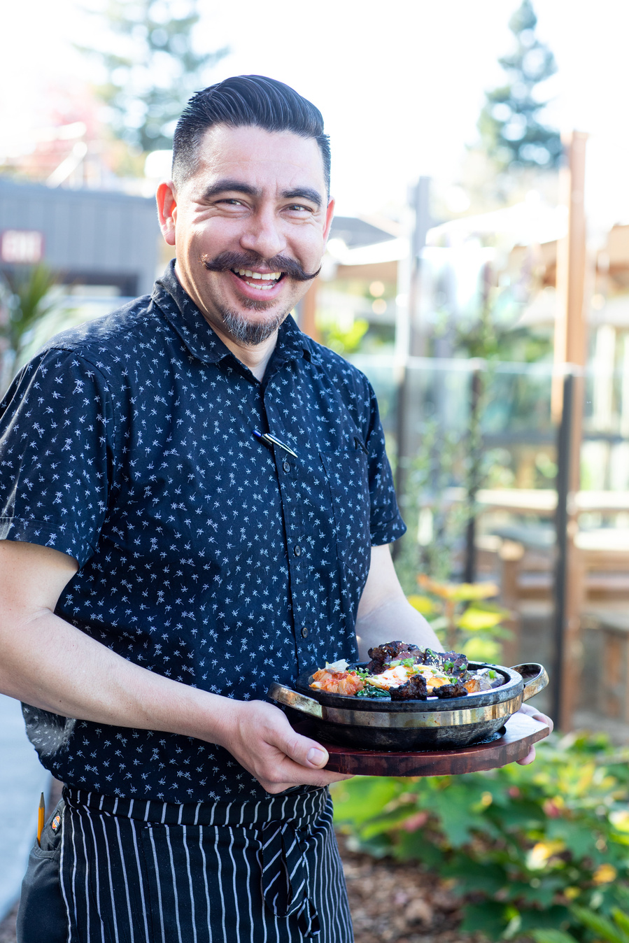 Smiling chef with mustache in black and white outfit, holding a steaming dish of food  on the outdoor patio of the restaurant. Flamingo resort restaurant photography. 