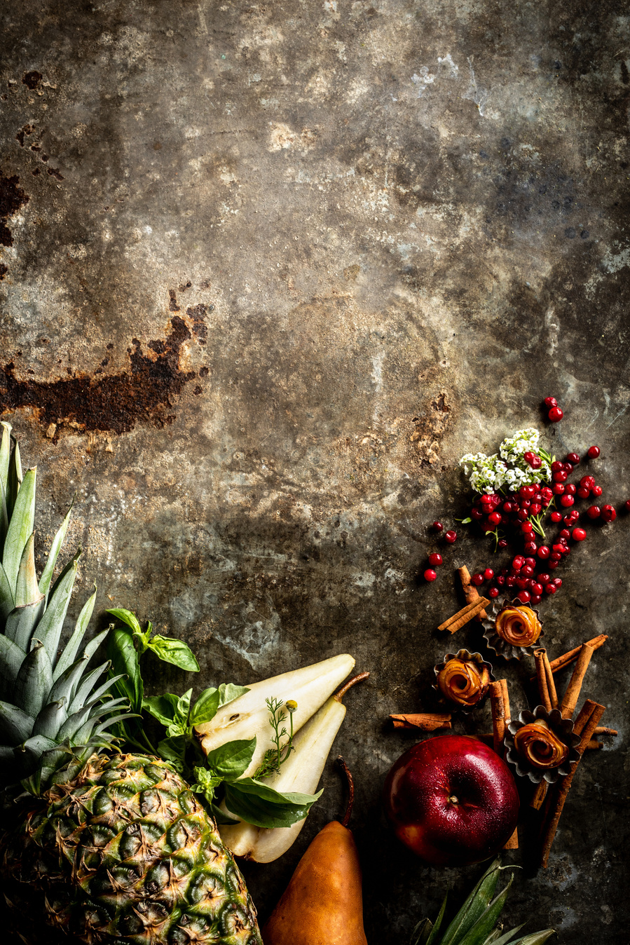 Overhead flat lay of pineapple, basil, apple, cinnamon, lingonberry, elderflower, pear, and cardamom on the bottom half of a rusted metal surface by a drink photographer  