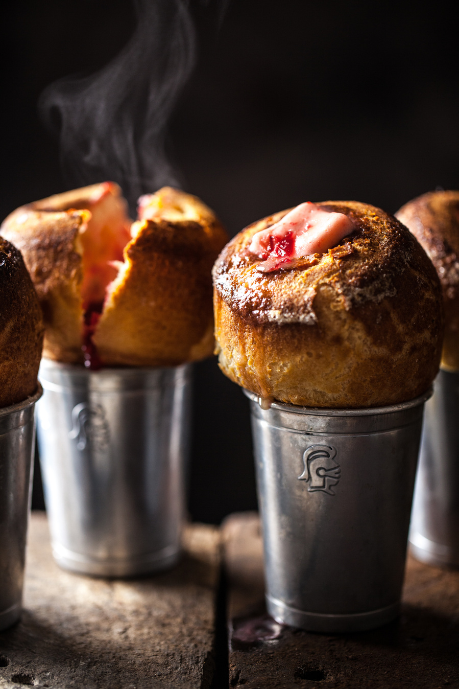 Dark food photography of strawberry popovers spilling out of shiny tins and steaming. Hard light highlights the baking photography