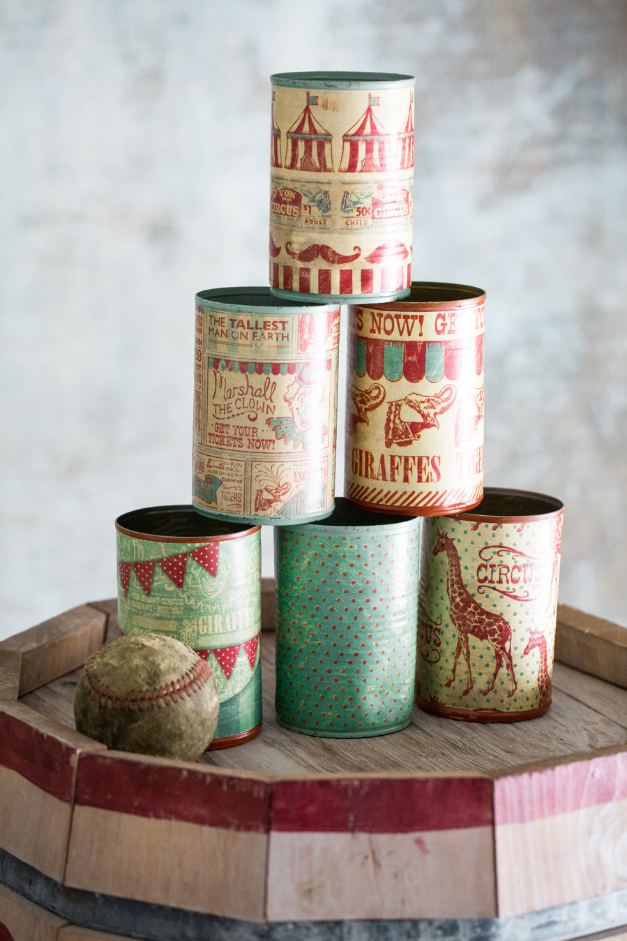 Tower of cans stacked on a barrel covered in vintage paper with a baseball for a DIY carnival game. Part of a circus themed birthday party. 