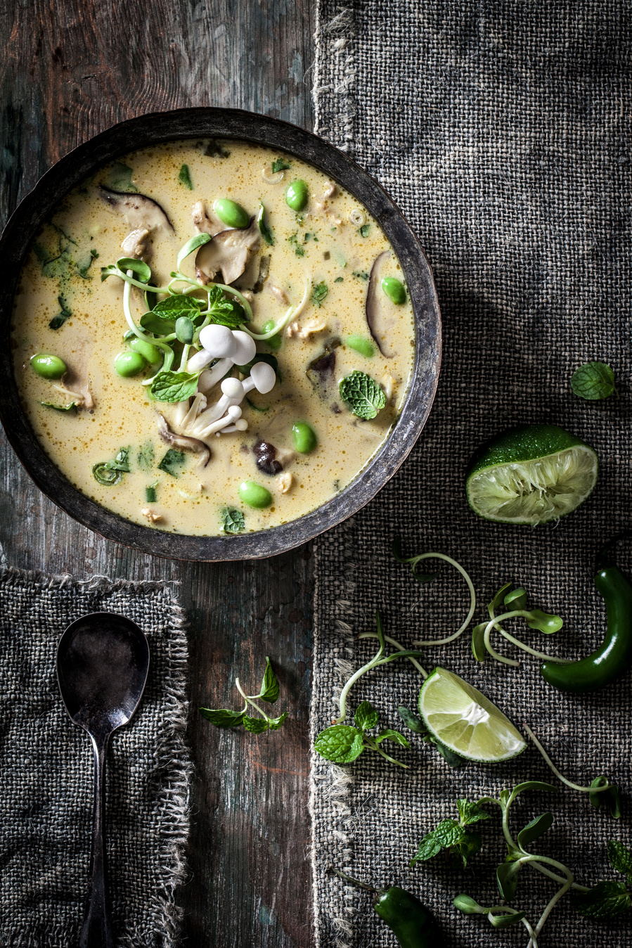 Overhead food photography bowl of mushroom soup. Dark food photography surface and props with yellow soup, and limes and greens on the surfaces. 