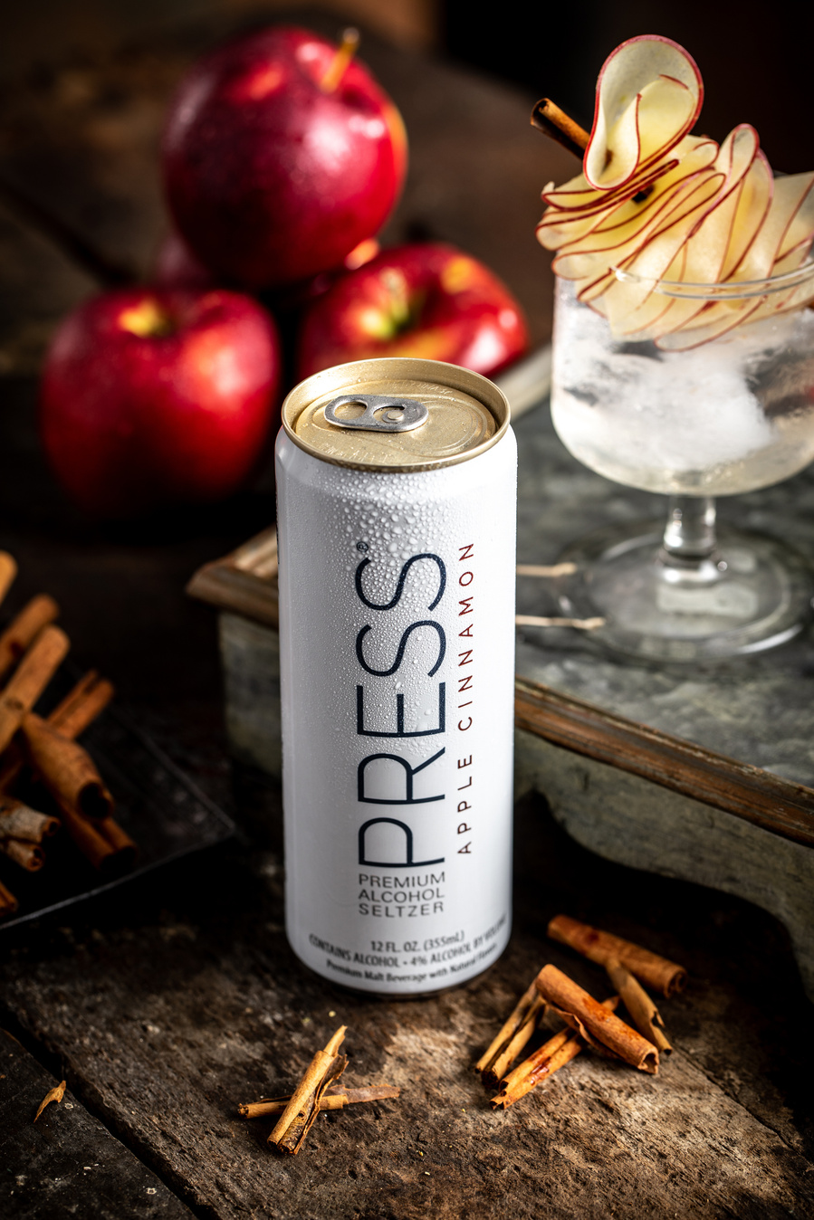 Can of apple cinnamon press seltzer with garnished drink  and apples behind and cinnamon sticks on the surface. 