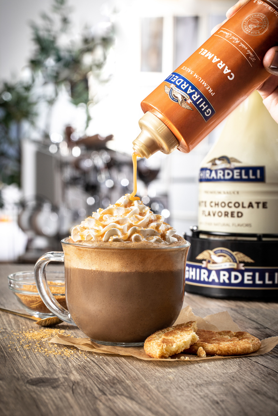 Big glass mug with a latte topped with whipped cream on top and Ghirardelli caramel sauce being added on top. Ghirardelli chocolate creamer package in background.