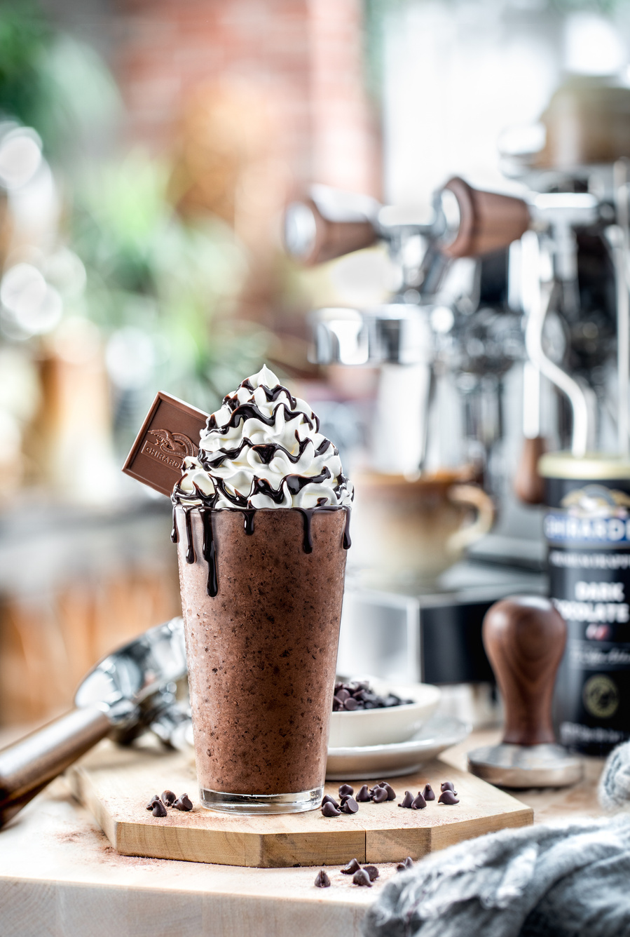 Frozen chocolate mocha with whipped cream, chocolate sauce, and Ghirardelli square in a tall glass in front of a shiny espresso machine.  Bright artistic chocolate photographer style. 